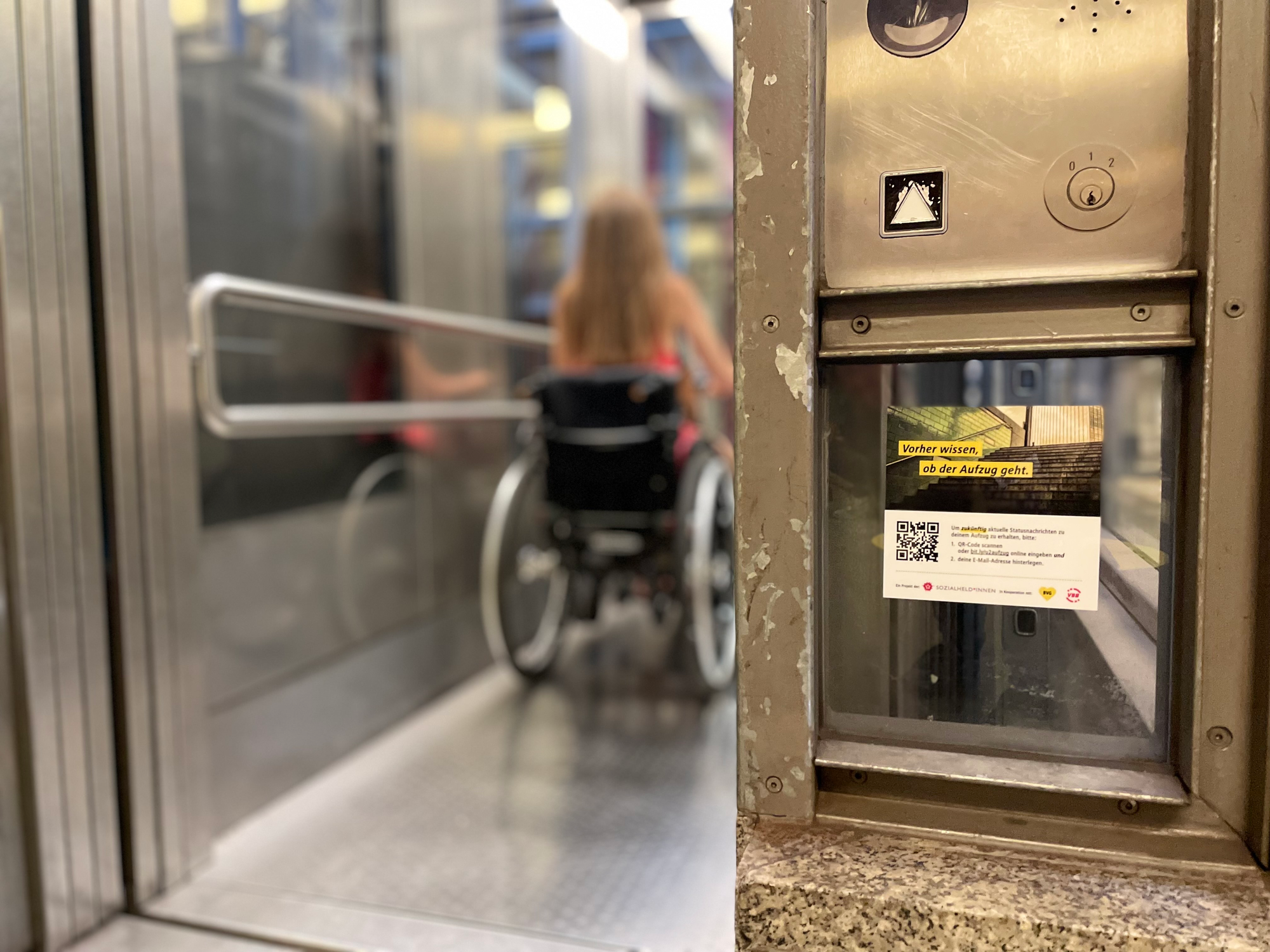 A person using a wheelchair right after entering an elevator. Right under the elevator’s controls, a sticker with a QR code advertises that you can get notifications from this elevator if there is a service disruption.